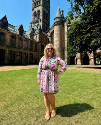 A woman in a colorful floral dress and sunglasses poses on a sunlit lawn in front of a historic Gothic Revival university building with a tall stone tower, turret, and arched cloisters