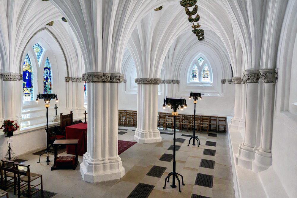 Bright Gothic cathedral interior with white ribbed vaulted ceilings, clustered stone columns, stained-glass windows, decorative ceiling bosses, chairs, and freestanding lights.
