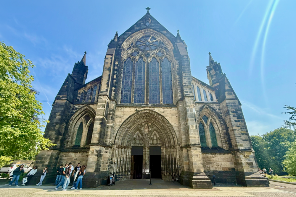 Front facade of a Gothic Revival cathedral featuring a large pointed-arch entrance, ornate stone tracery, tall stained-glass windows, and twin spires beneath a bright blue sky.