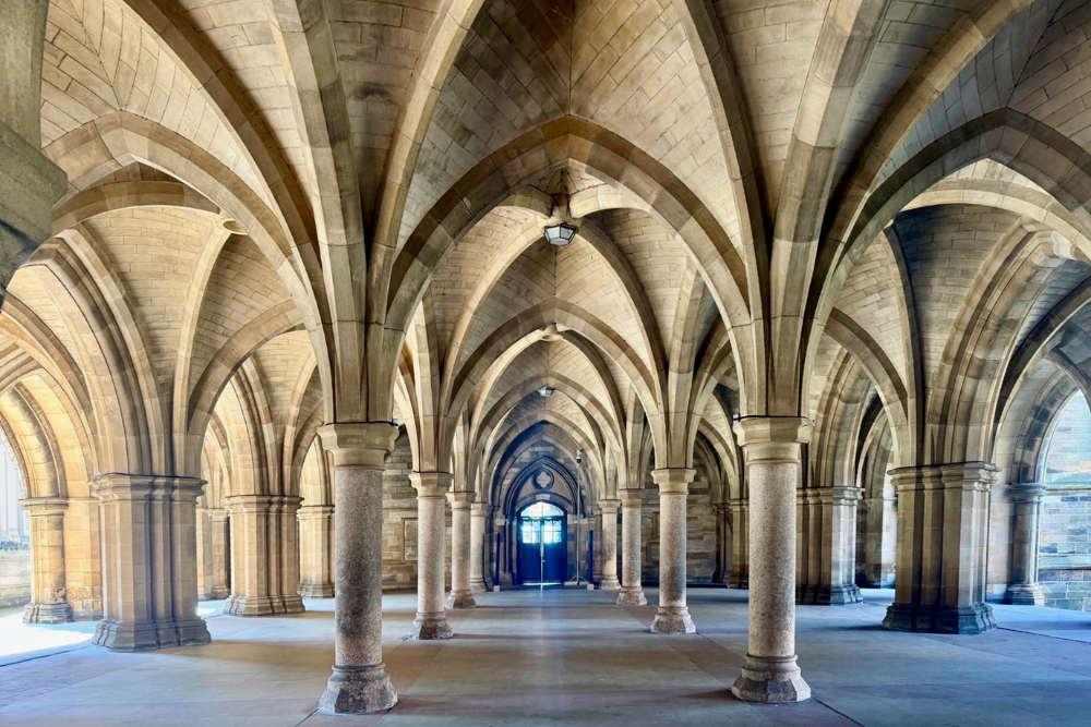 Interior of a Gothic Revival stone cloister featuring symmetrical pointed arches, ribbed vaulted ceilings, and rows of stone columns extending toward a central doorway