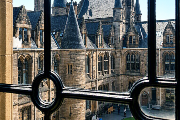 View through a decorative black-framed window overlooking a historic Gothic Revival university courtyard with stone turrets, steep slate roofs, pointed arches, and ornate windows