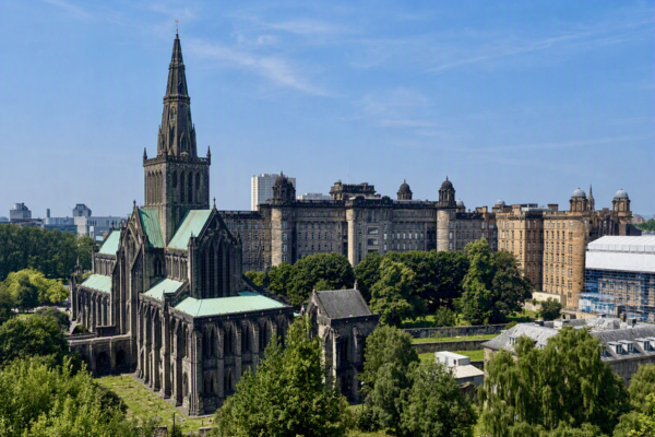 Glasgow Cathedral and its tall Gothic spire rise above surrounding trees, with the historic Glasgow Royal Infirmary complex visible in the background beneath a clear blue sky.