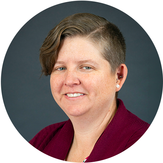 Professional headshot of a smiling woman with short asymmetrical brown hair, earrings, and a burgundy cardigan against a dark gray background