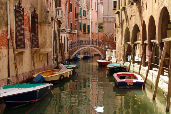 Narrow Venice canal lined with historic buildings, small moored boats, and an arched pedestrian bridge reflected in the water