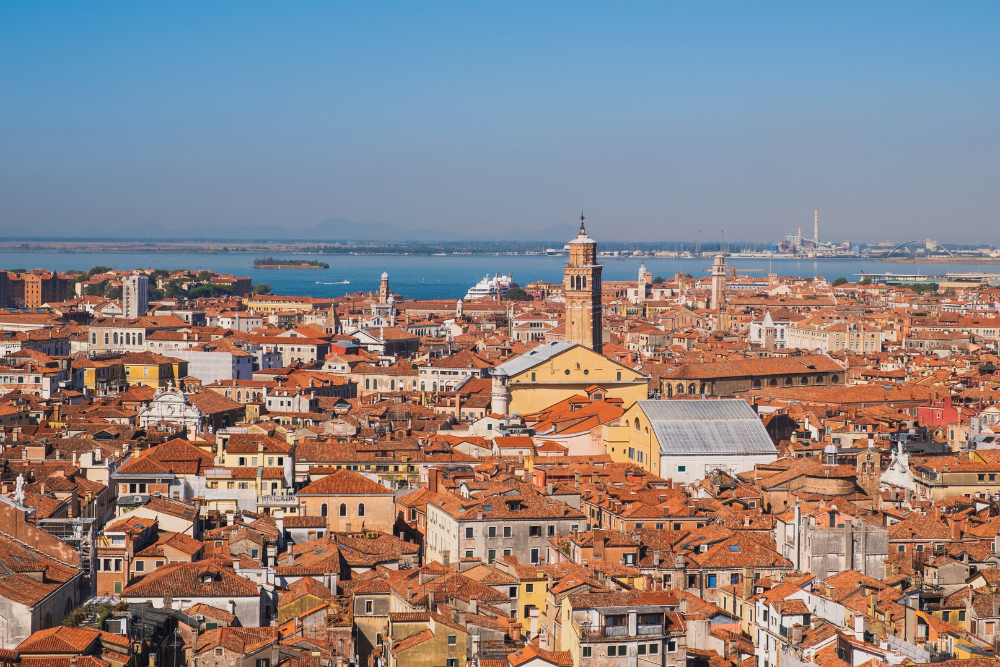Panoramic view of Venice, Italy, showing dense terracotta rooftops, historic churches and bell towers, with the Venetian Lagoon beyond