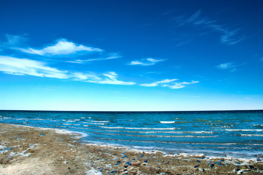 Shallow blue waters of the Aral Sea meeting a sandy, salt-encrusted shoreline beneath a vivid blue sky with wispy white clouds