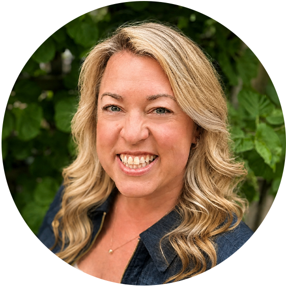 Professional headshot of a smiling woman with long wavy blonde hair wearing a dark blue jacket, photographed outdoors against a leafy green background