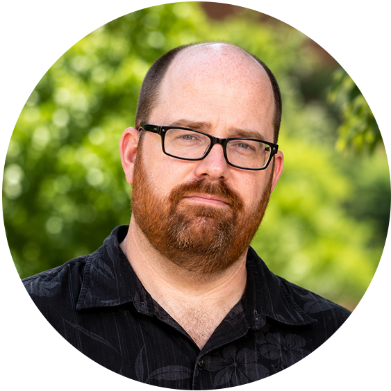 Professional headshot of a man with a reddish-brown beard, black rectangular glasses, and a black collared shirt against a blurred green outdoor background