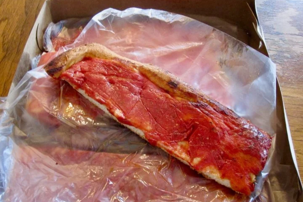 Traditional Rhode Island bakery pizza strip topped with thick red tomato sauce, resting on wax paper inside a cardboard takeout box